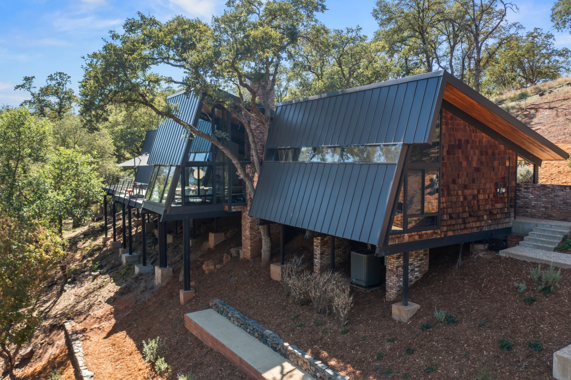 Low aerial of the home's angular standing-seam roofs and cedar-shingle siding among the oaks