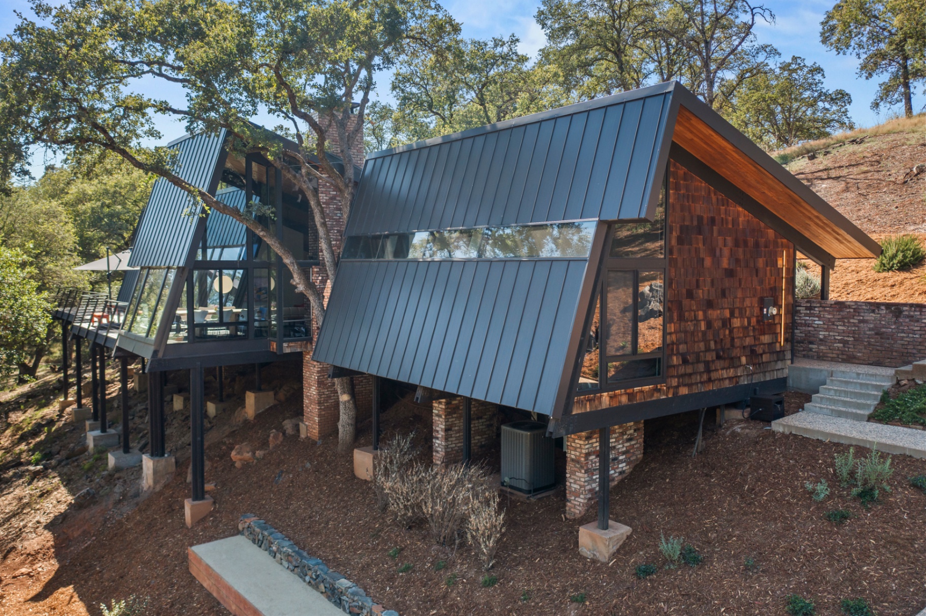 Side aerial of the home elevated on steel posts above the sloping hillside